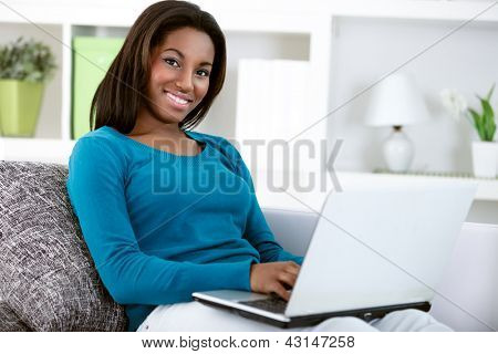 Picture or Photo of Young black girl with laptop computer, sitting in bright living room, looking at camera.