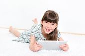 picture of laughing  - Joyful pretty little girl laughing as she lies on her stomach on a white carpet playing with a modern apple ipad in a white room - JPG 