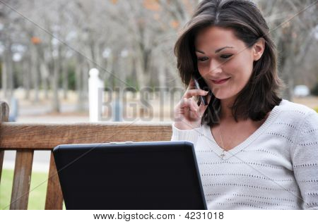 Picture or Photo of Young woman sitting on bench in park talking on cell phone and using laptop.