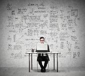 picture of economy  - Businessman working at a laptop with in the background a wall full of economy drawings - JPG 