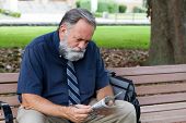 stock photo of newspaper  - Unemployed middle aged man looks at advertisements for jobs in a newspaper while sitting on a park bench - JPG 