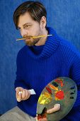 Artist in blue sweater adds oil paints on a palette. He holds bristles in his mouth.